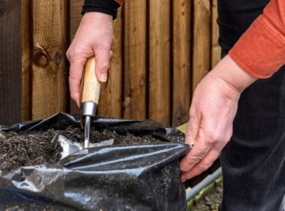 A person digging through bagged soil risks, using a trowel to work dark brown material inside a black plastic bag