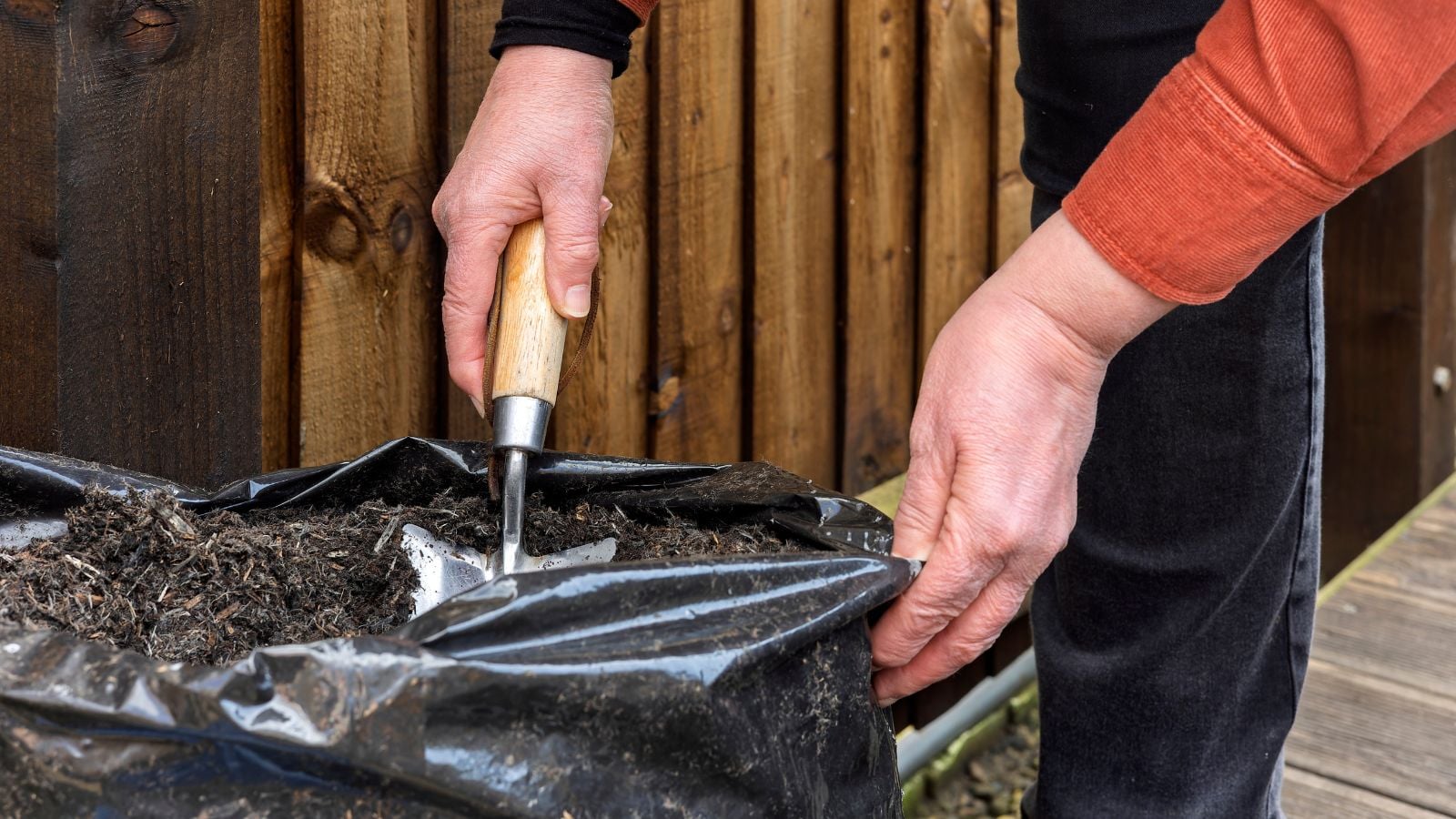 A person digging through bagged soil risks, using a trowel to work dark brown material inside a black plastic bag