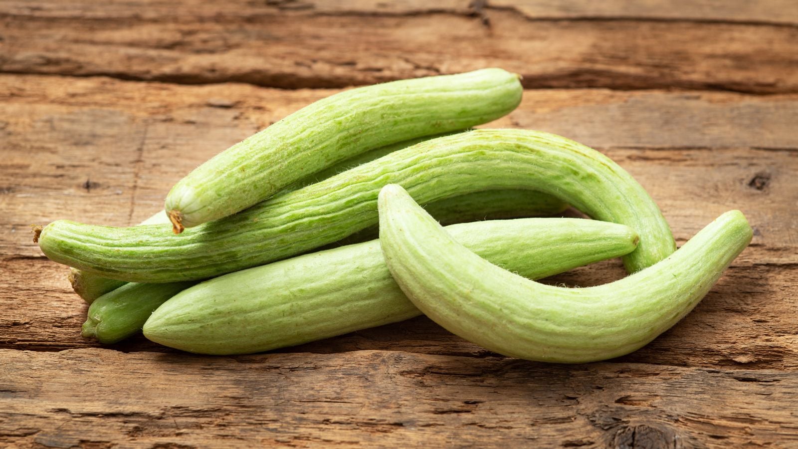 A close-up and overhead shot of slender, long, and slightly ribbed, light-green colored fruits of the Armenian melon, placed on a wooden surface