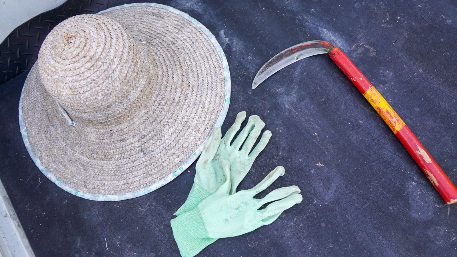 A close-up and overhead shot of a flat-lay arrangement of several equipment, including a straw hat, gloves and a sickle